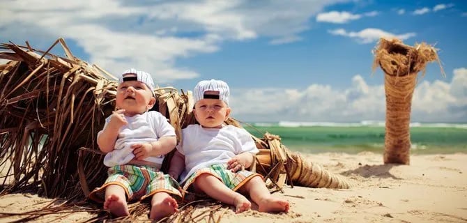 Kumsalda güneş şapkalı iki bebek yan yana oturuyor