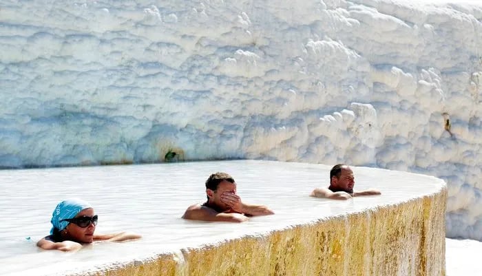 Pamukkale’de bir traverten havuzunda suya girmiş üç ziyaretçi; arkada beyaz teras duvarı