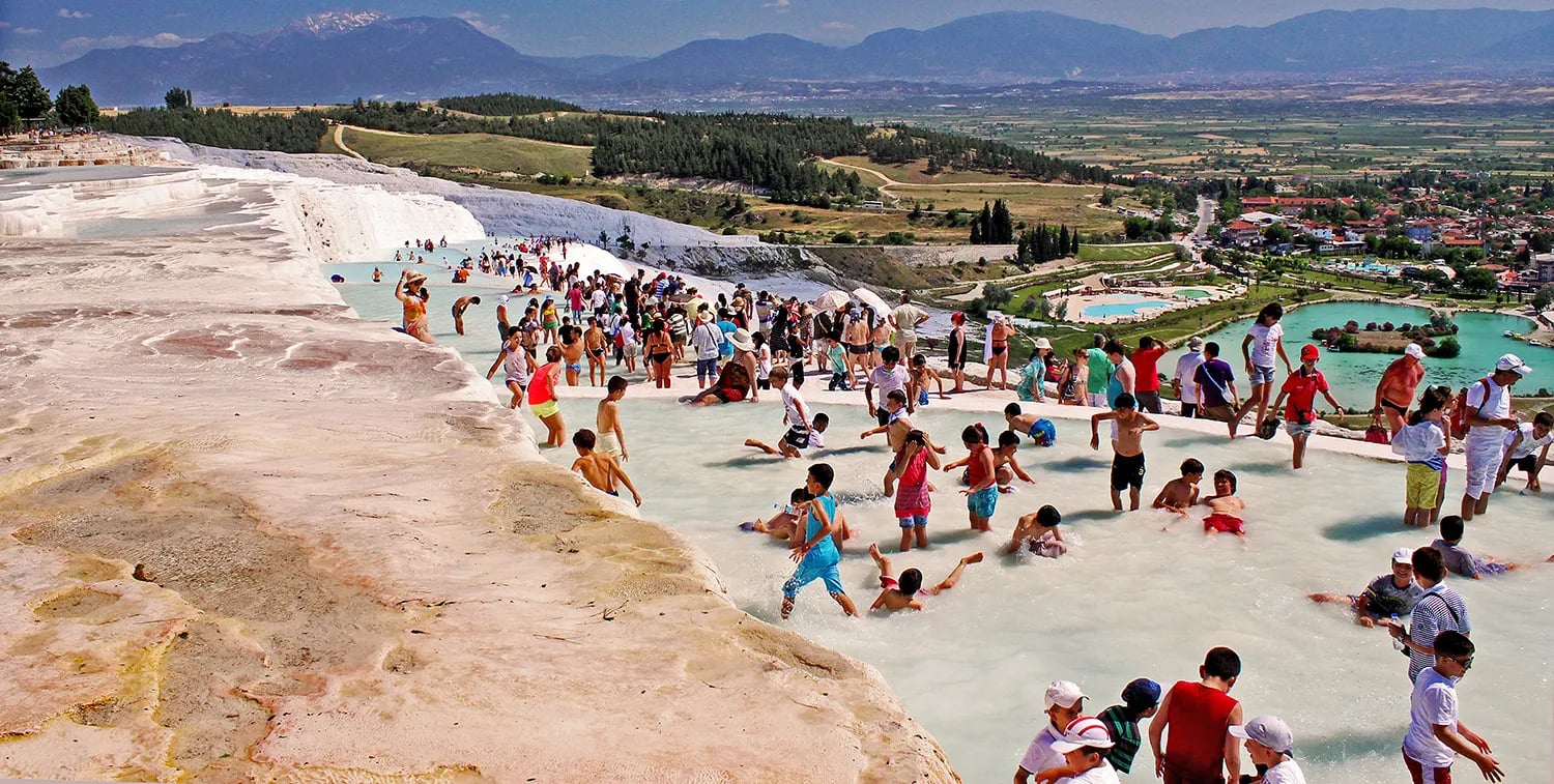 Pamukkale travertenlerinin tepesinden geniş görünüm: turkuaz teras havuzlarında serinleyen ziyaretçiler, arkada Denizli ovası