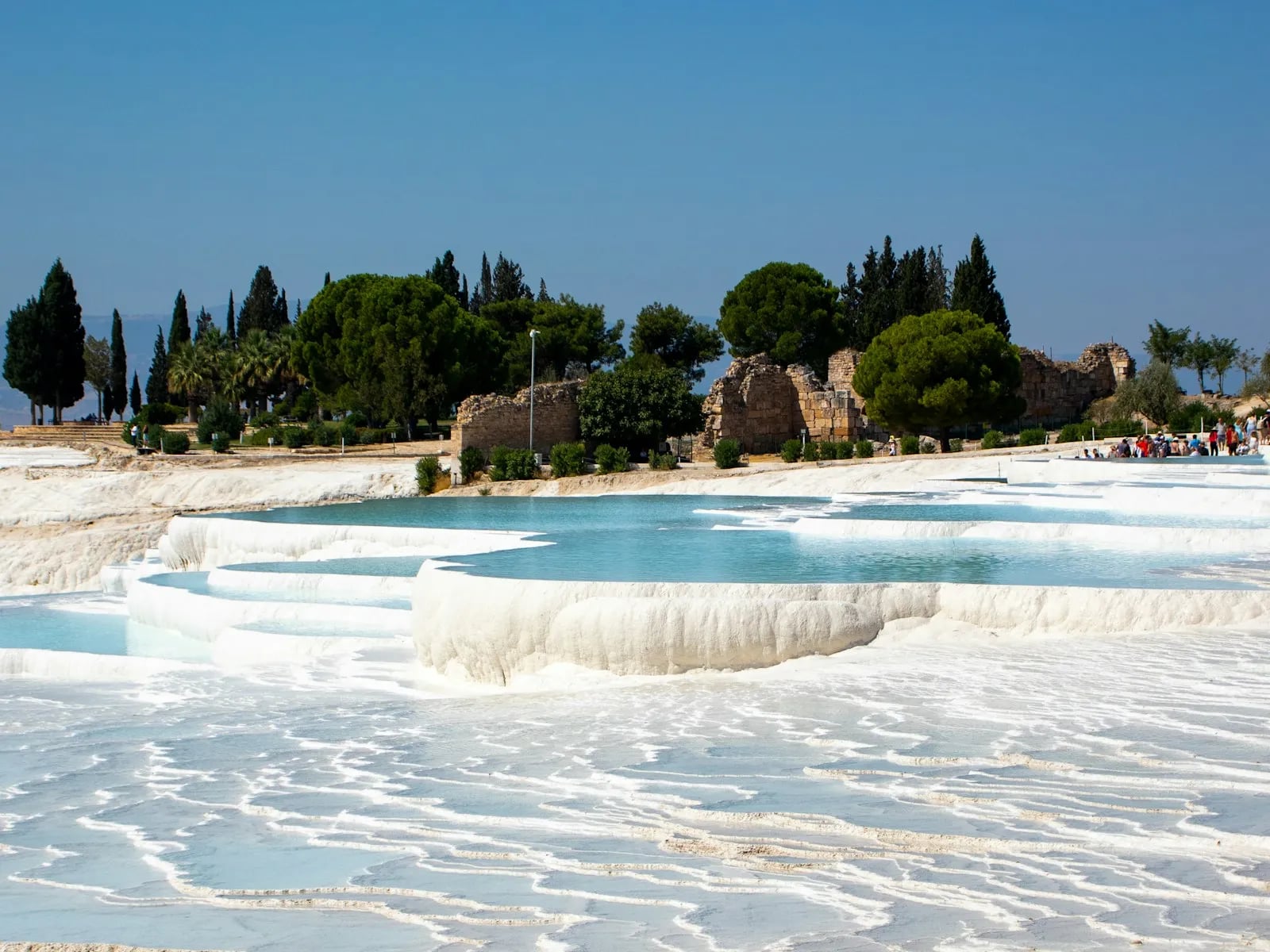 Pamukkale’de boş traverten terasları ve turkuaz sığ havuzlar; arka planda Hierapolis kalıntıları ile selvi ve çam ağaçları