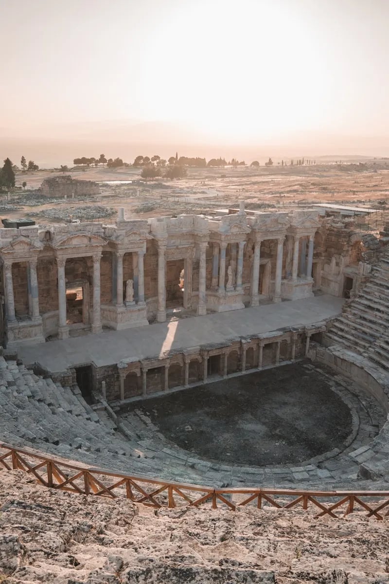 Hierapolis Antik Tiyatrosu’na yukarıdan bakış: yarım daire oturma sıraları, sahne binası ve puslu ışıkta uzanan Denizli ovası