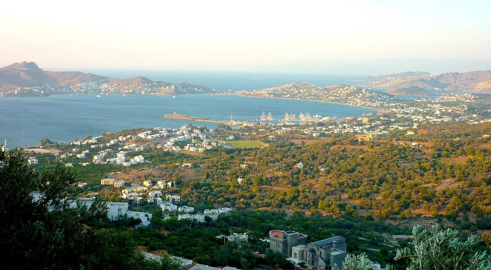 Tepeden Yalıkavak Körfezi: altın ışıkta uzanan koy, marina, zeytinlikler ve beyaz köy dokusu