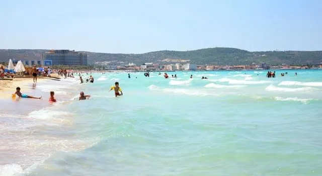 Çeşme'de geniş kumsal ve turkuaz sığ su; kıyıda denize giren insanlar, arkada tesisler ve alçak tepeler