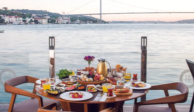 Boğaz kenarında kurulmuş zengin kahvaltı sofrası; arkada köprü ve karşı kıyı