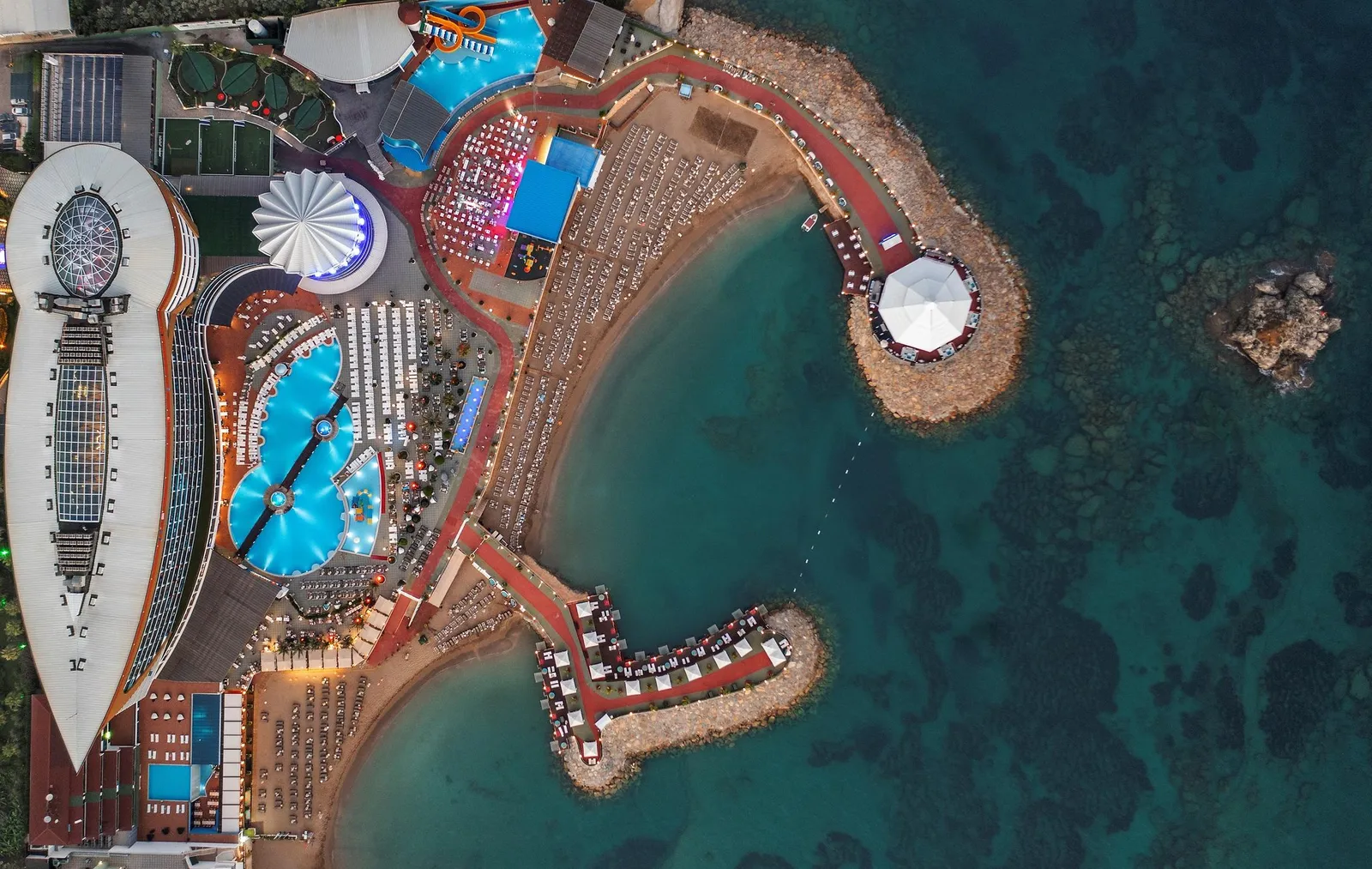 Otelin kuş bakışı gece görünümü: aydınlatılmış havuzlar, plaj ve deniz üstü yuvarlak platform