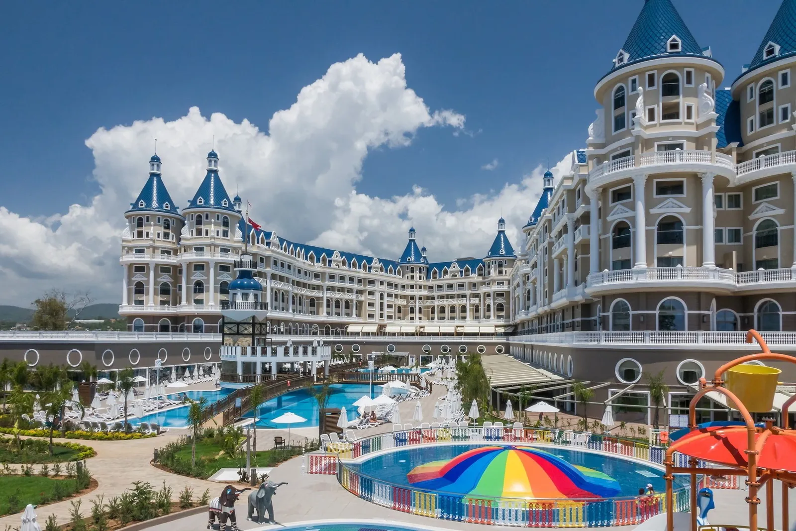 Kuleli, saray görünümlü otel cephesi; önde gökkuşağı şemsiyeli çocuk havuzu